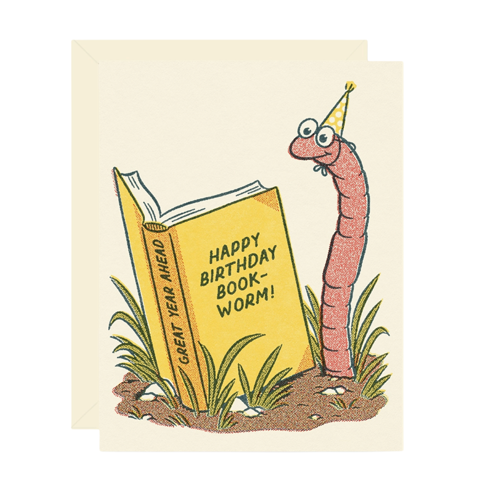 A greeting card with a cream envelope behind it, featuring a whimsical illustration of a pink worm wearing a yellow party hat beside an open yellow book that reads “HAPPY BIRTHDAY BOOK-WORM!” and “GREAT YEAR AHEAD.”.