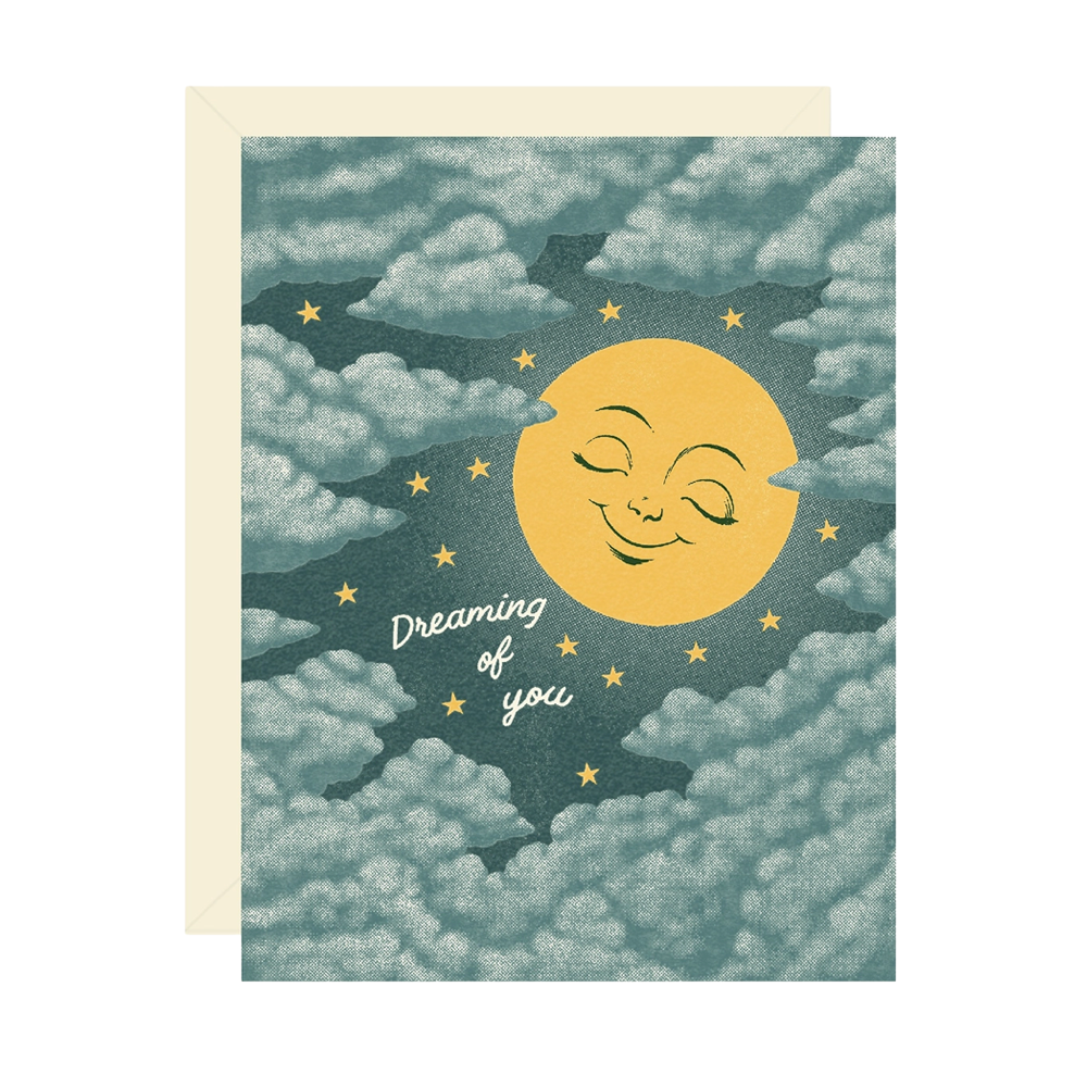 A greeting card with a whimsical celestial design featuring a smiling yellow sun and moon among clouds, with the words 'Dreaming of you' in white script. The card is shown with a cream envelope behind it on a white background.