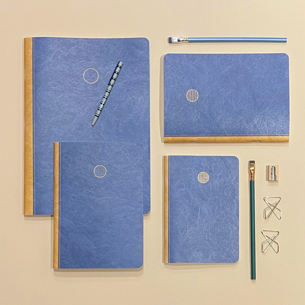 A coordinated stationery set arranged on a warm beige background, including three blue textured notebooks with tan edges, a blue pencil, a blue eraser, a paintbrush, a small metal sharpener, and metal paperclips.