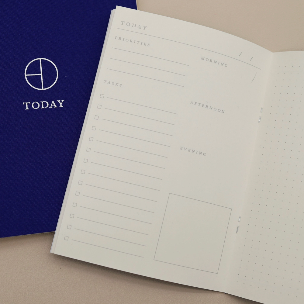 An open planner showing white pages with minimalist daily planning sections and a dotted grid, placed next to a dark blue cover printed with the word TODAY and a circular logo.