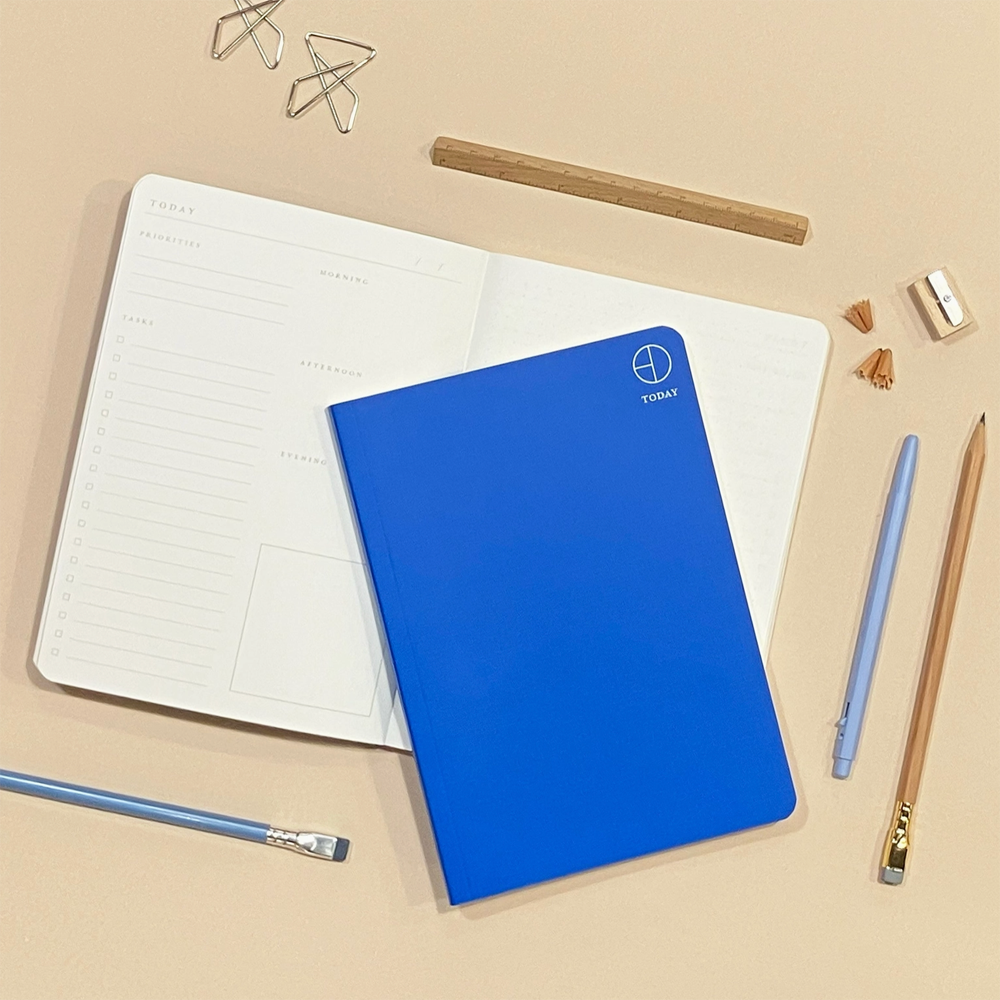 A bright blue TODAY planner notebook with a white logo, placed on top of an open white daily planner. The scene is styled as a flat lay on a beige surface with a wooden pencil, a light blue pen, a wooden pencil, metal paperclips, and a small eraser.