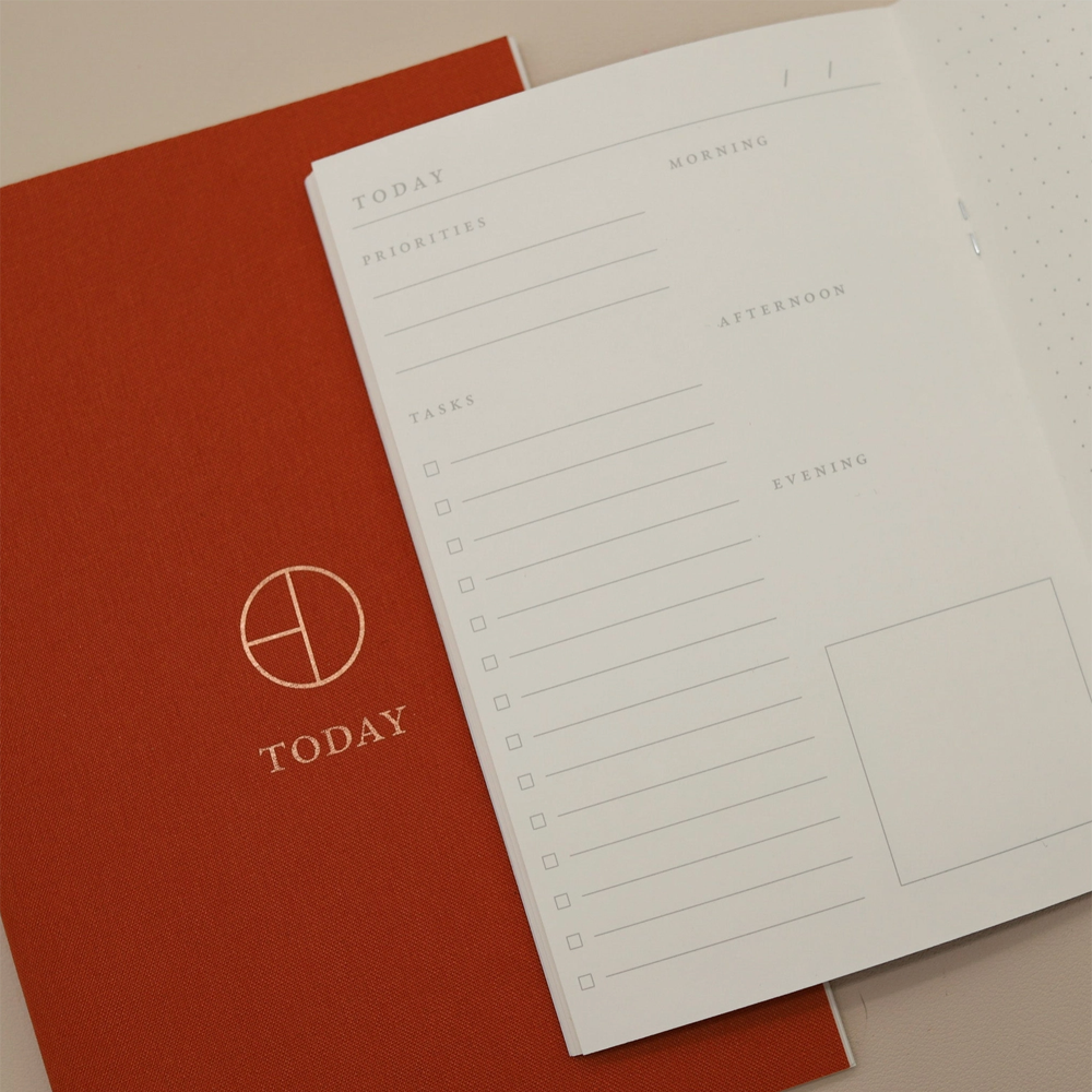 An open white planner showing a daily planner page with sections for today, priorities, tasks, and times of day, placed over a rust-red cover with a gold circular logo and the word TODAY.