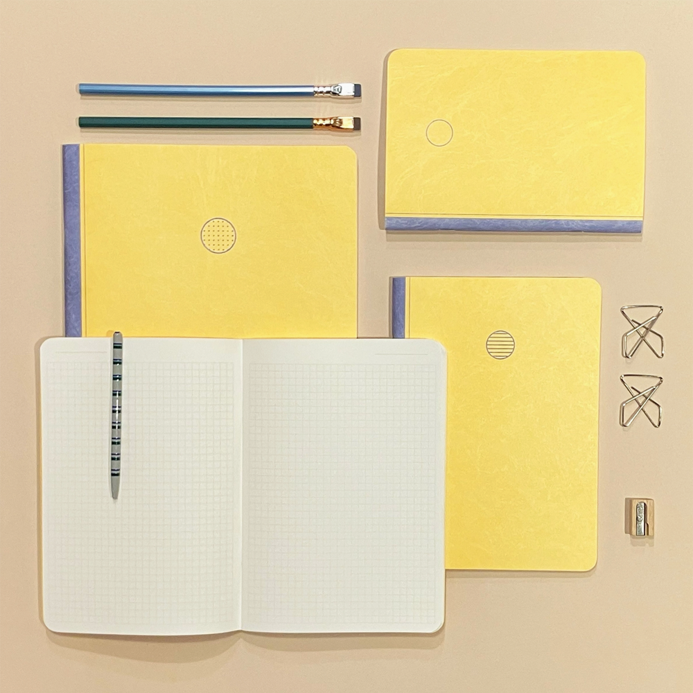 A flat-lay arrangement of stationery items on a warm beige background, including three yellow notebooks, an open white grid notebook, a pen, two pencils, two metal paperclips, and a pencil sharpener.