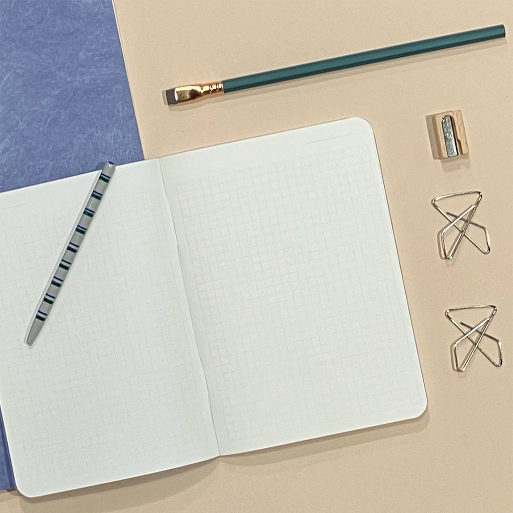An open grid notebook on a beige surface with a blue textured corner, styled with a teal pencil, a striped pen, a wooden eraser, and two metal paperclips.