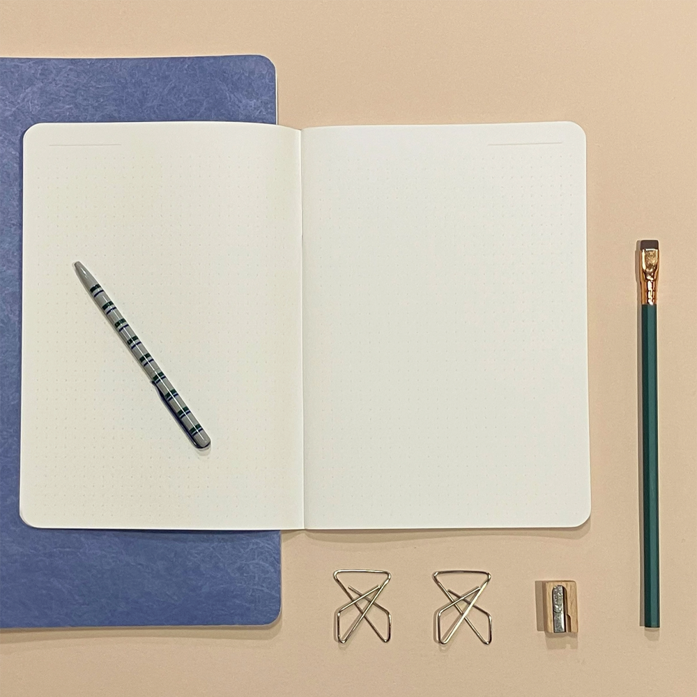 An open notebook with blank white pages displayed on a beige background, alongside a blue textured cover, a striped pen, a green pencil, two metal paperclips, and a small pencil sharpener.