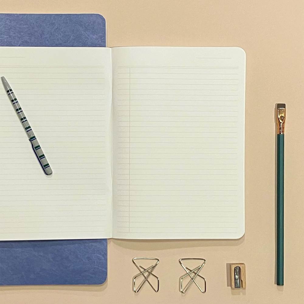 An open lined notebook with a blue cover lies on a beige surface, styled with a striped pen, a green pencil, two metal binder clips, and a small wooden eraser.