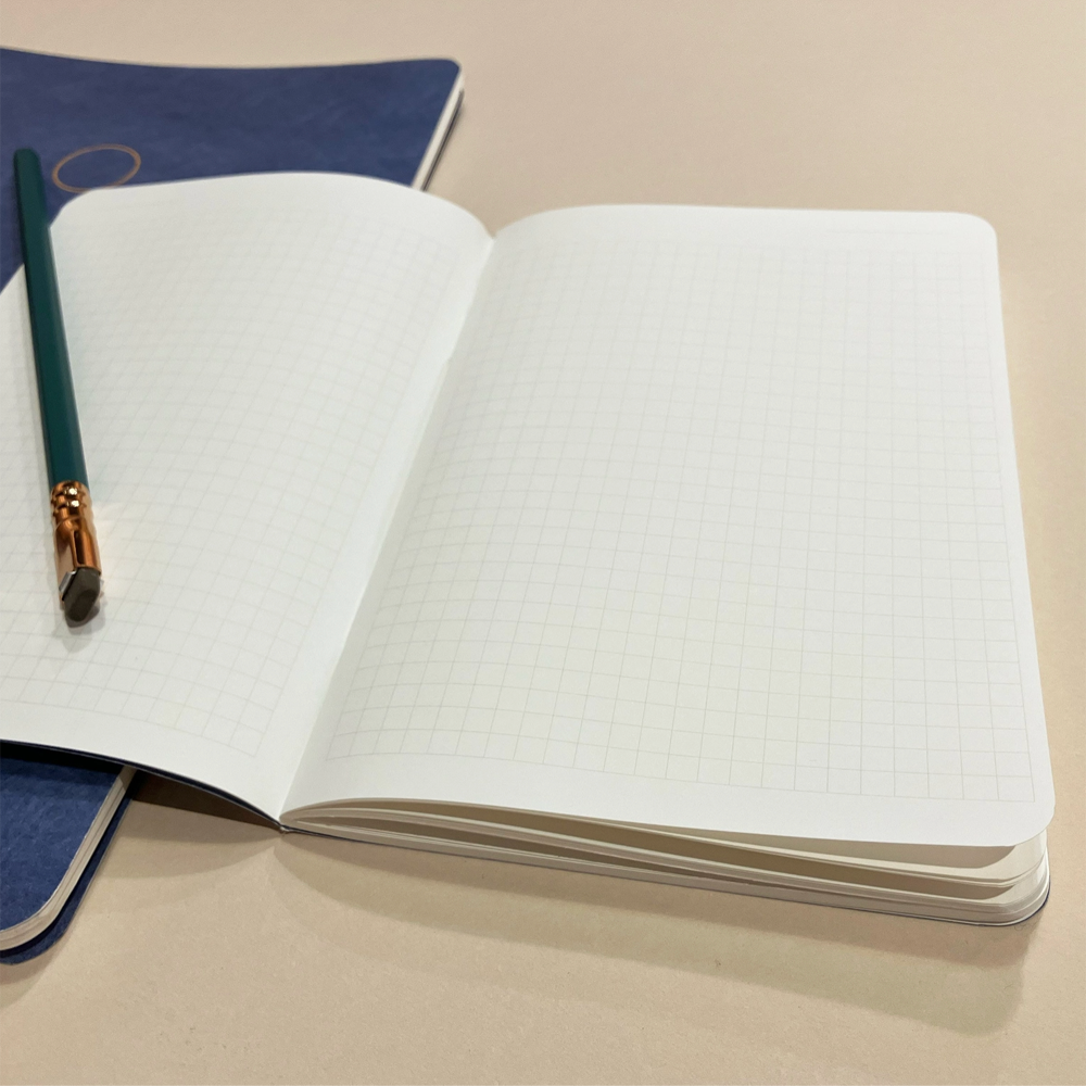 An open notebook with white grid paper pages, photographed on a light beige surface. A dark blue closed copy of the same notebook and a teal pencil with a gold ferrule are placed beside it.
