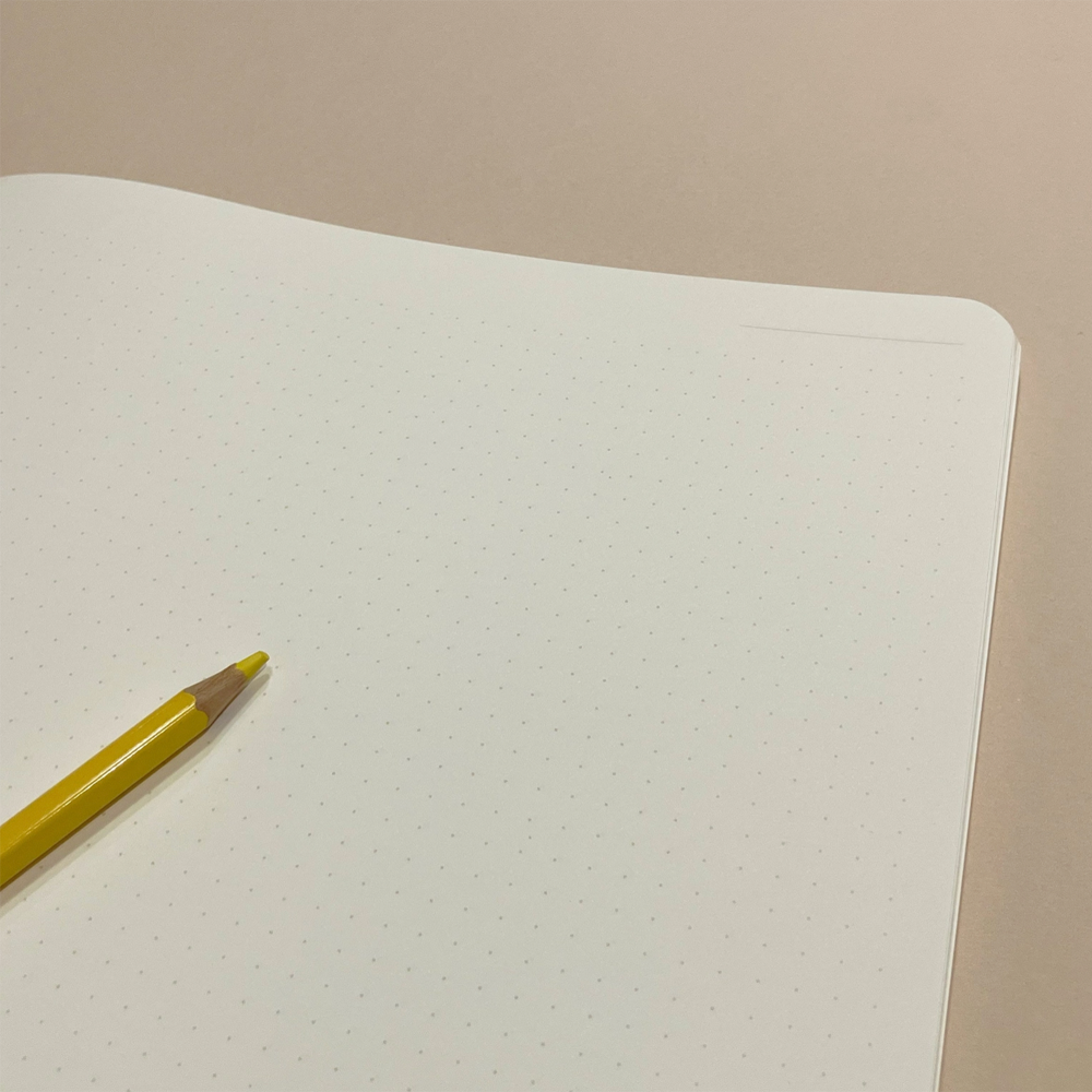 A close-up of a white dot grid notebook page with a yellow pencil resting on it against a warm beige background.