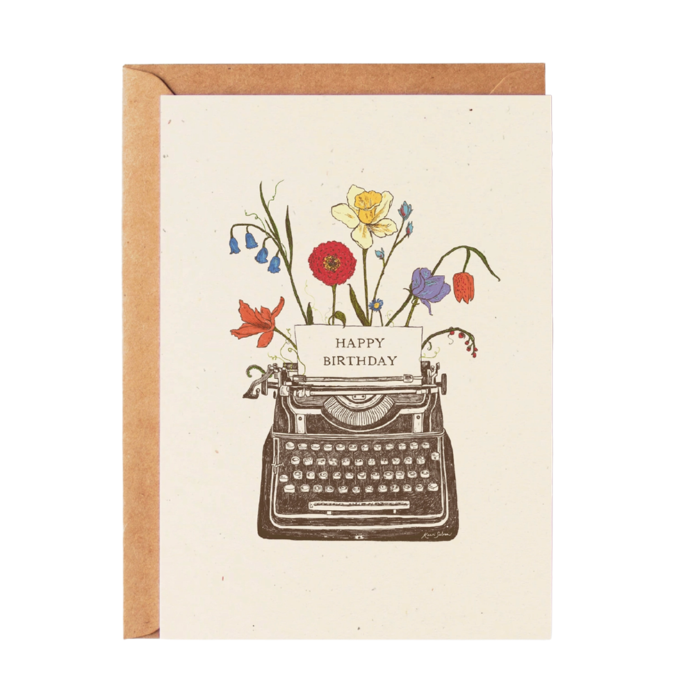 A greeting card with a kraft envelope, featuring an illustration of a vintage typewriter with colorful wildflowers and a card reading 'HAPPY BIRTHDAY' on a light cream background.