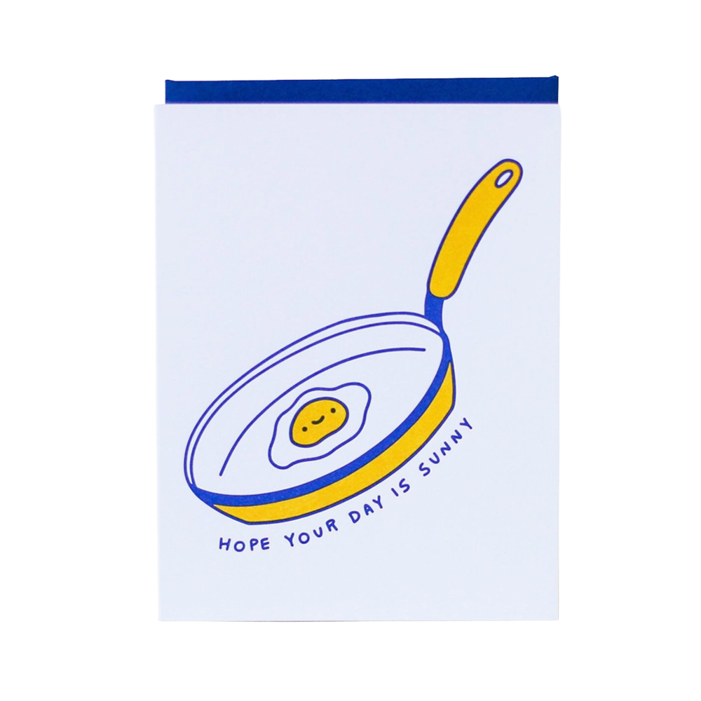 A white greeting card featuring a hand-drawn illustration of a frying pan with a smiling yellow egg inside, with the text 'HOPE YOUR DAY IS SUNNY' written below in blue. The card is shown with a dark blue envelope or backing at the top against a plain white background.