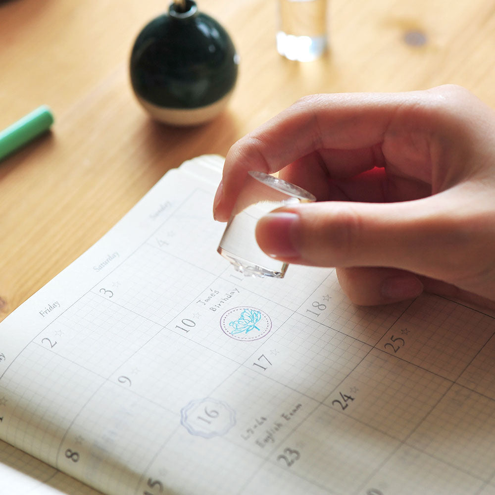 Hand using a small acrylic clear cylinder on a calendar with a pen and decorative item in the background.
