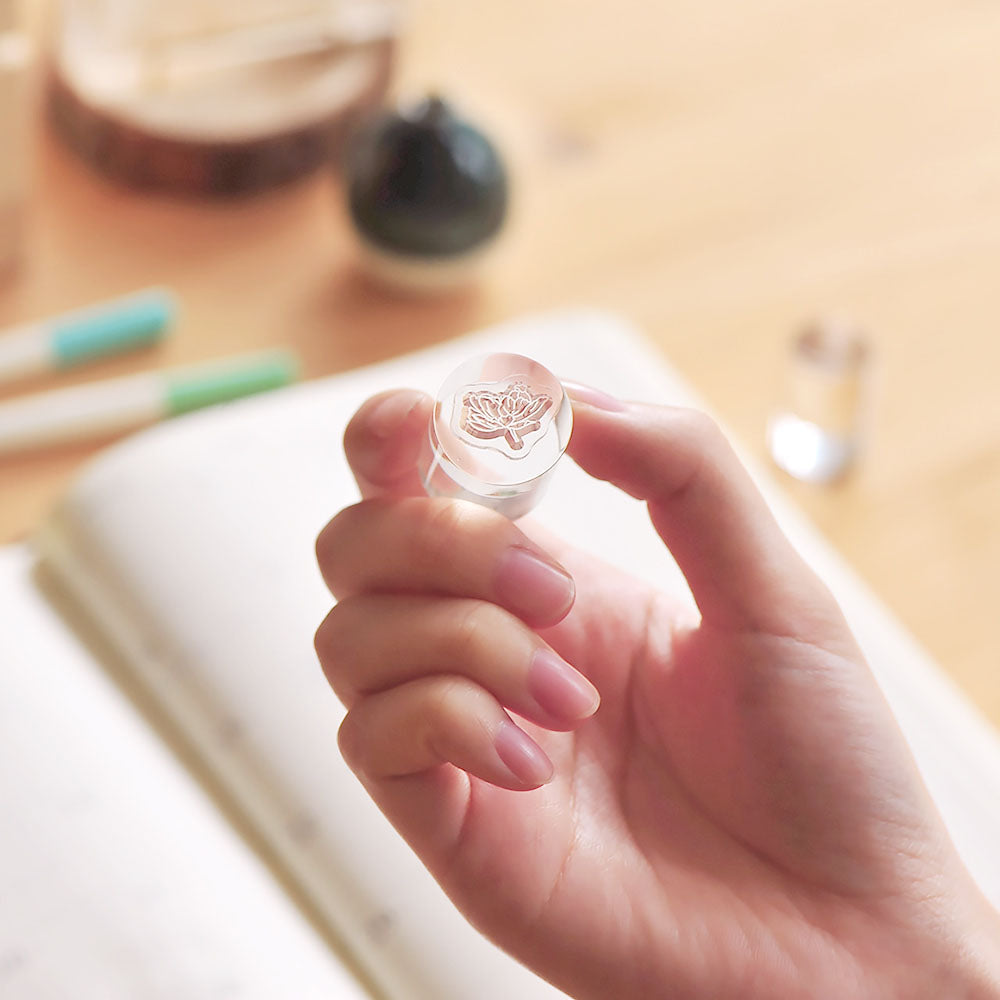 Hand holding a small clear cylinder with a stamp on it on a background of a desk with stationery items.