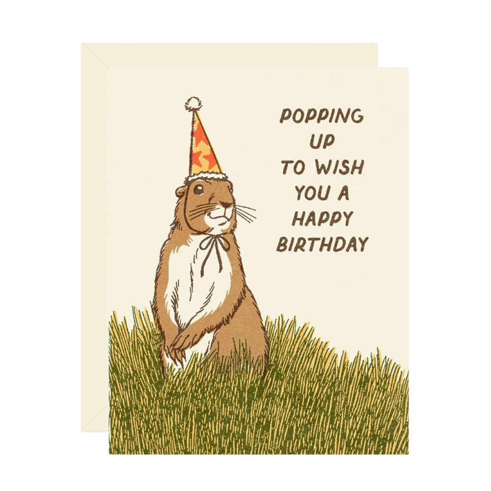 A greeting card with a cream background featuring an illustration of a brown prarie dog wearing a red party hat in green grass, next to the text 'POPPING UP TO WISH YOU A HAPPY BIRTHDAY'