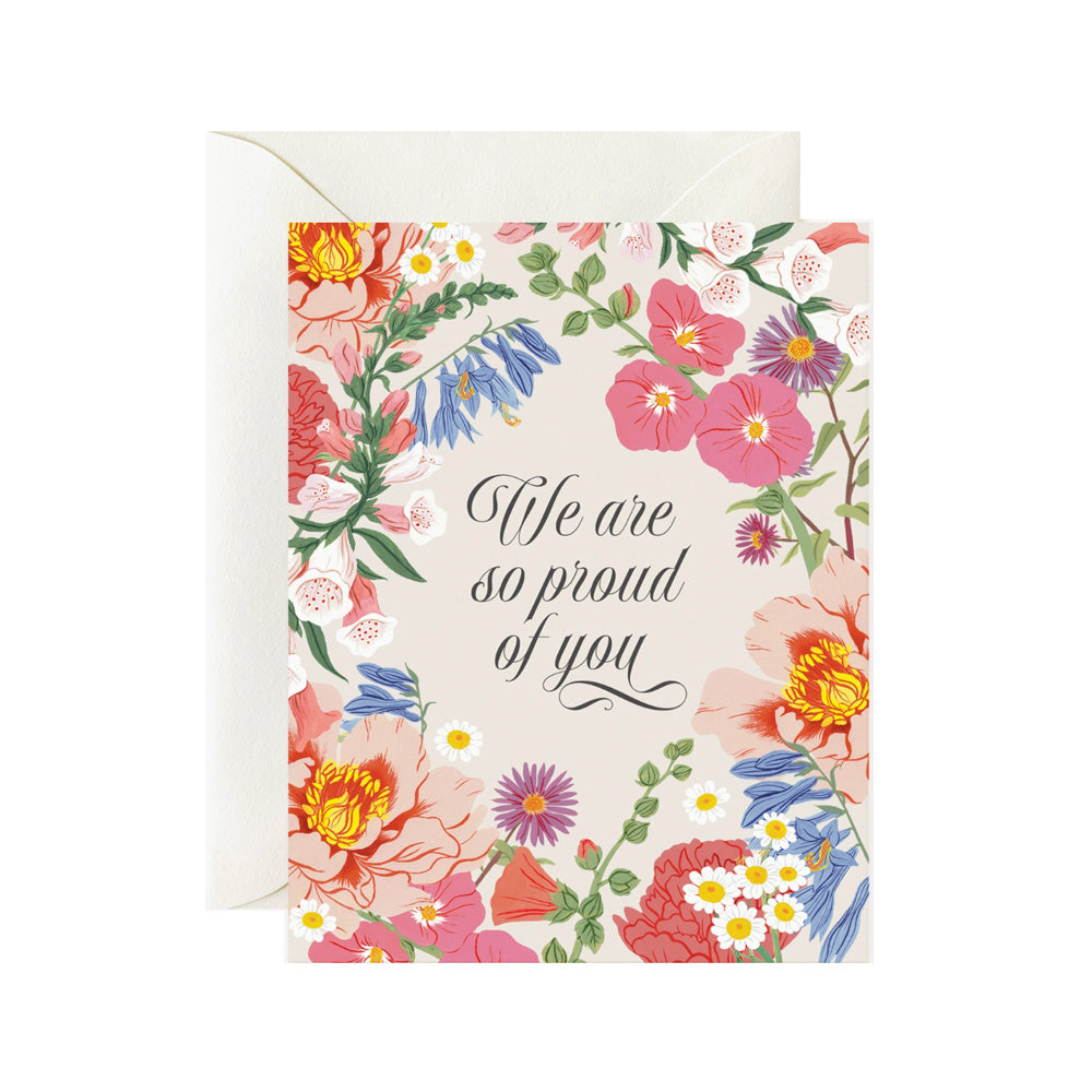 A cream color card with multiple flowers along the edges with the words "We are so proud of you" in the center in a script font.
