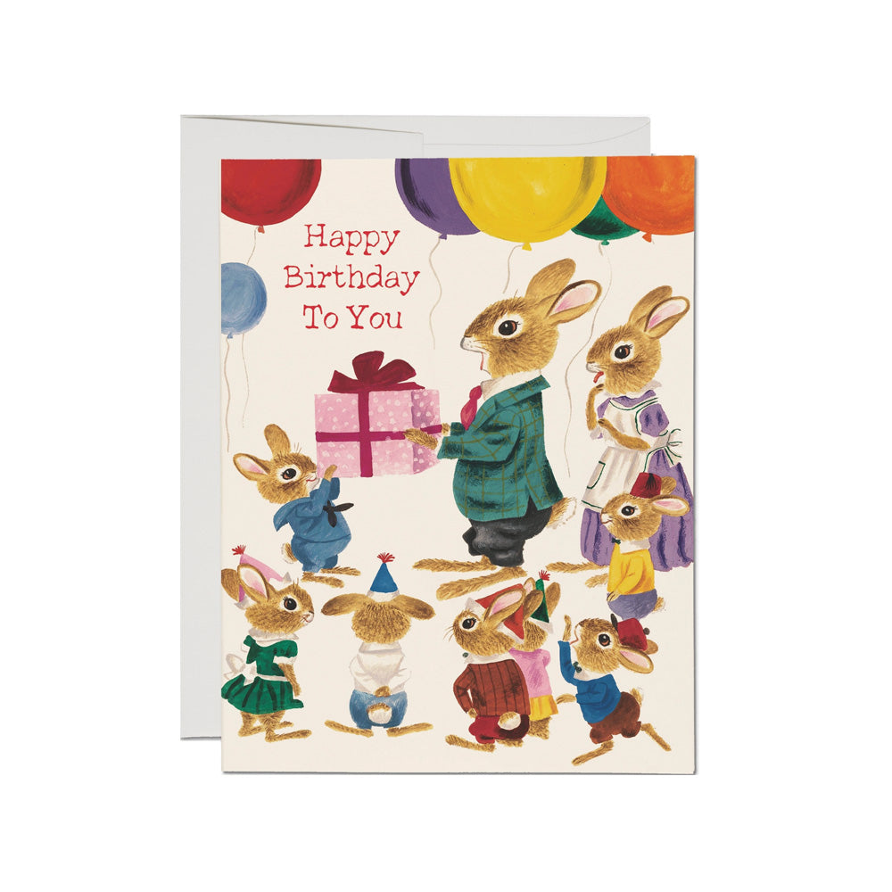 Birthday card with two parent bunnies and a bunch of kid bunnies celebrating with a present and balloons on a white background.Text reads "Happy Birthday To You"
