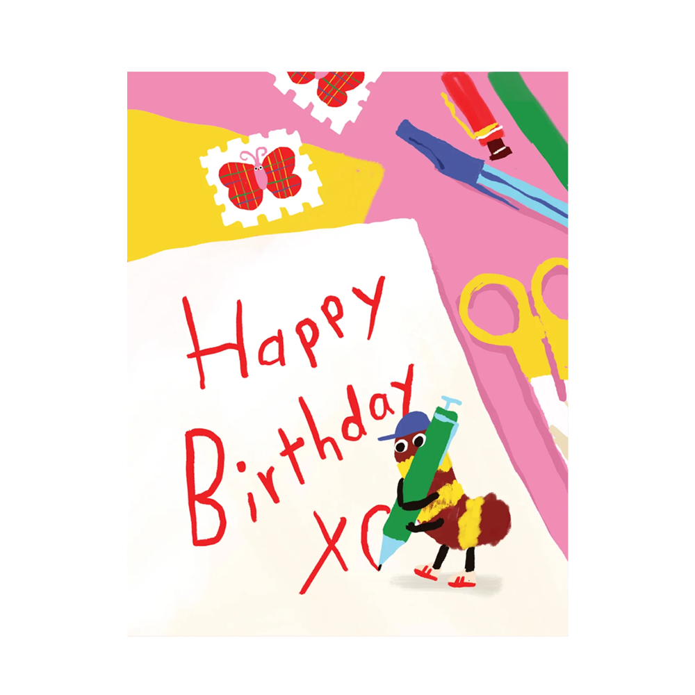 A colorful birthday card featuring the words 'Happy Birthday' in red script on a white sheet, surrounded by playful illustrated objects including scissors, crayons, stamps, and a small bug character holding a green pen.