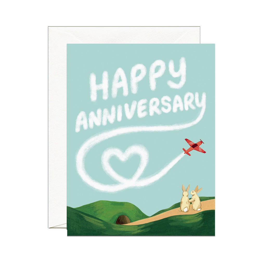 Greeting card with 'Happy Anniversary' text, heart-shaped smoke trail, and two rabbits on a path.