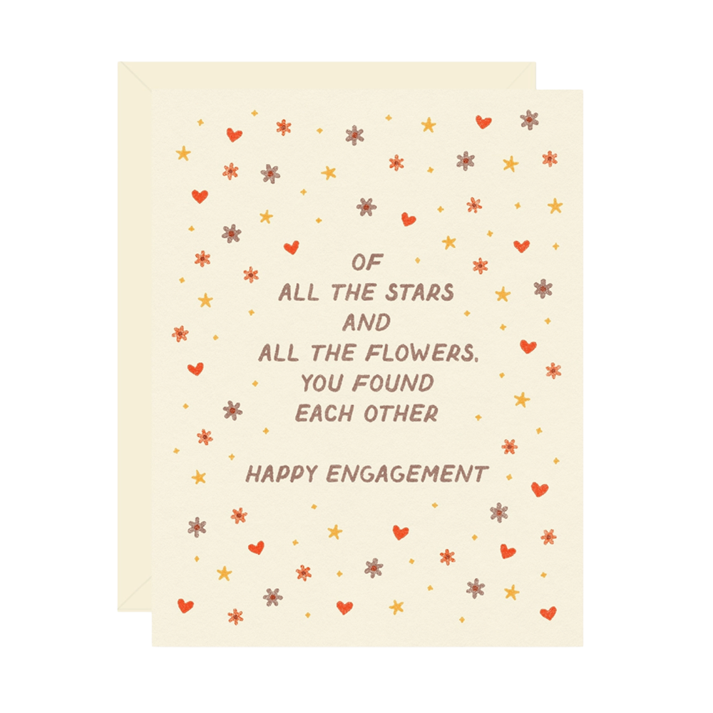 A cream-colored greeting card with a matching envelope, featuring a playful message that reads 'OF ALL THE STARS AND ALL THE FLOWERS, YOU FOUND EACH OTHER HAPPY ENGAGEMENT' surrounded by small scattered stars, hearts, and flowers in warm orange, brown, and gold tones.