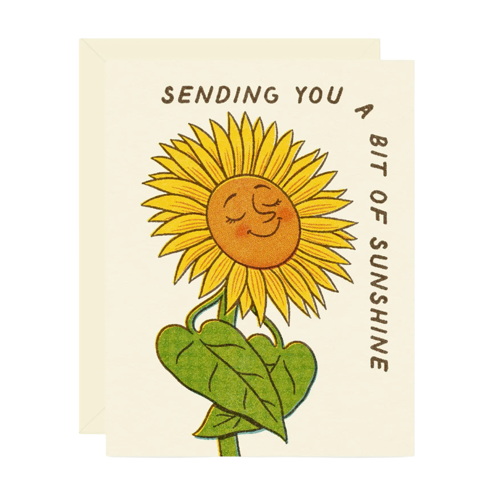 A cream-colored greeting card with a matching envelope, featuring a hand-drawn style illustration of a smiling sunflower with the text 'SENDING YOU A BIT OF SUNSHINE'.