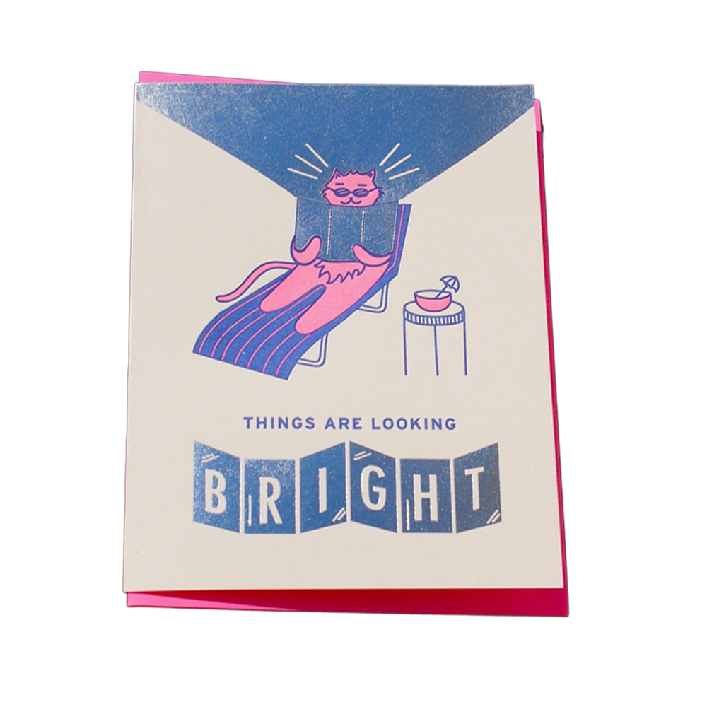 A greeting card with a cream-colored front, featuring a pink cat lounging on a blue striped chaise lounge under a blue spotlight, with the text 'THINGS ARE LOOKING BRIGHT' below. A bright pink envelope is visible behind the card on a white background.