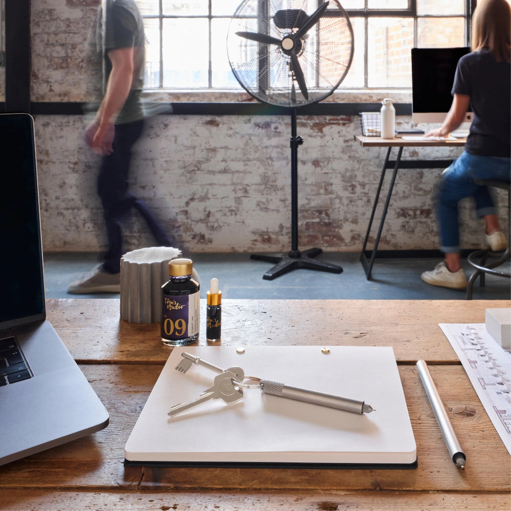 Desk with a notebook, pen, and bottles in an office setting with people working in the background.