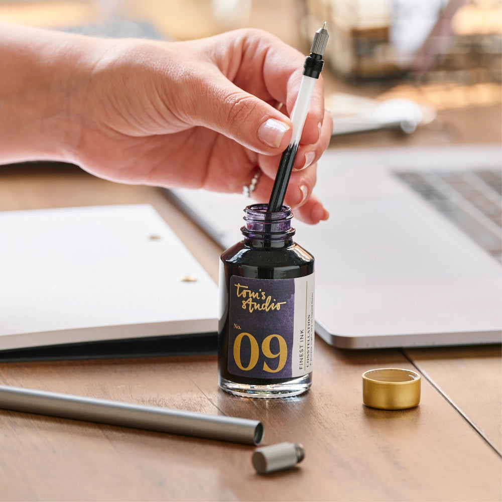 Person filling a pen with ink from a bottle labeled 'Toni's Studio 09' on a wooden desk.