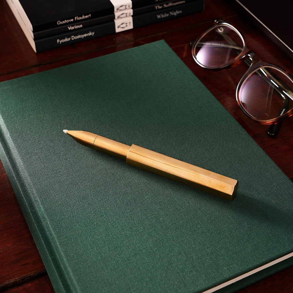 Green notebook with a Brass pen on a wooden surface, with books and glasses in the background.