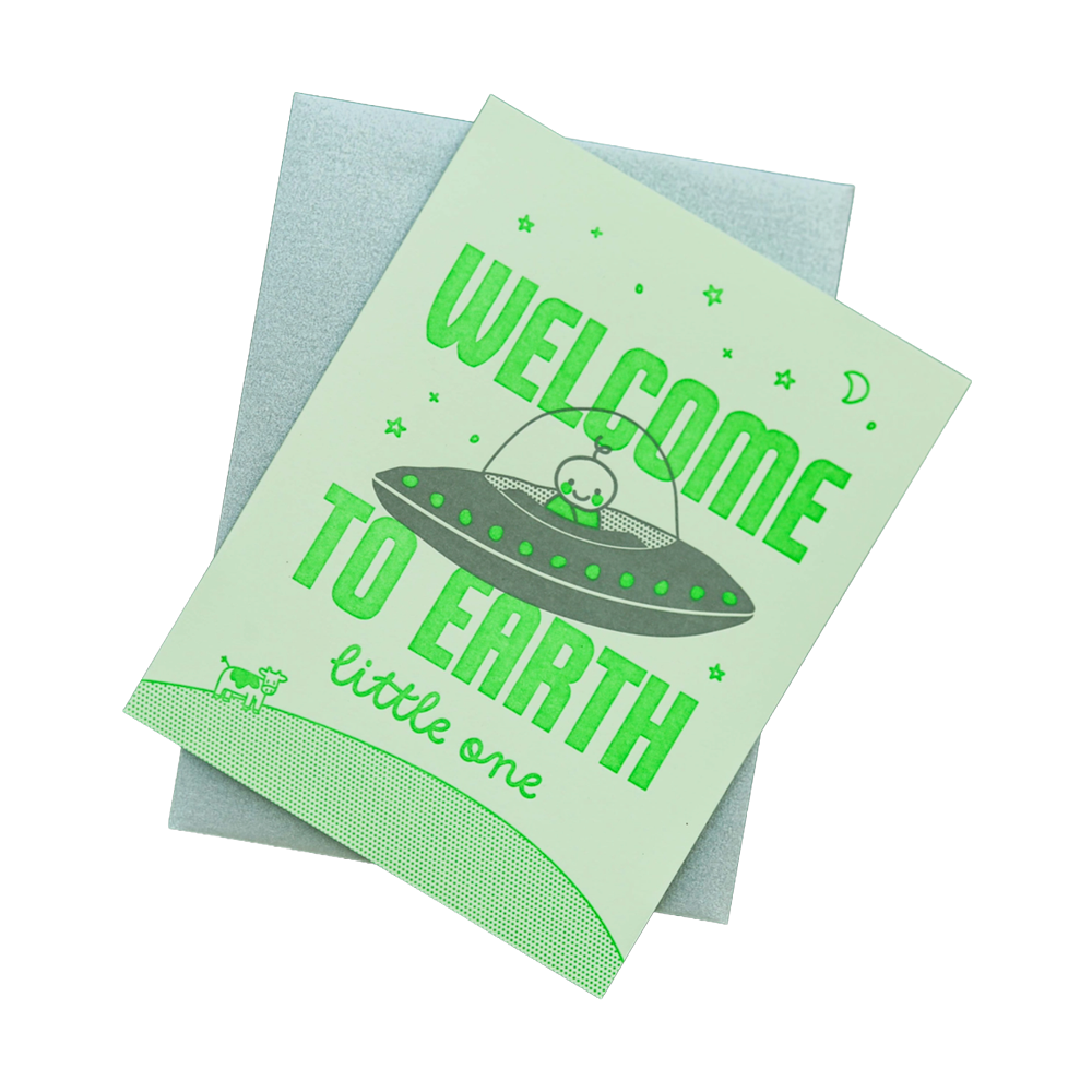 A light green greeting card with a matching light blue envelope, featuring a retro-style illustration of a UFO and the text 'WELCOME TO EARTH little one'.