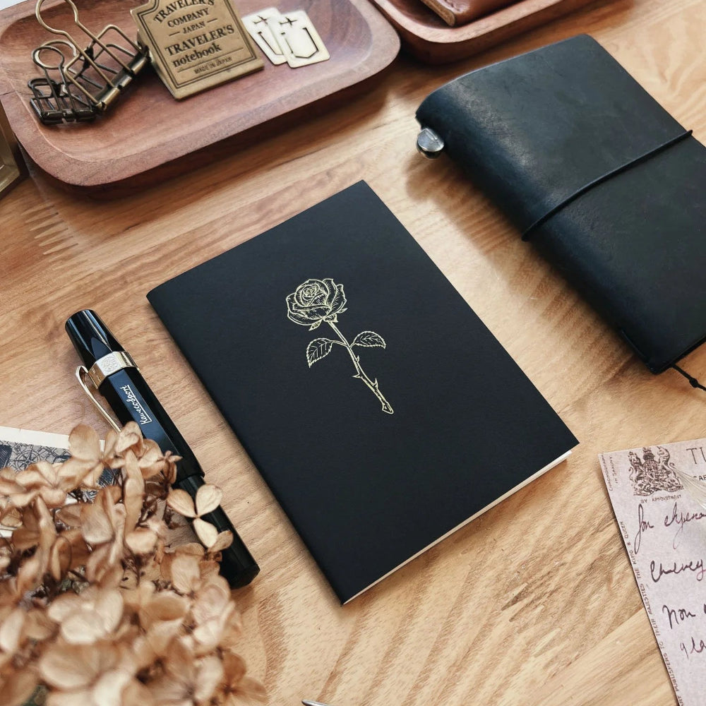 Black notebook with a rose design on a wooden surface with stationery items.