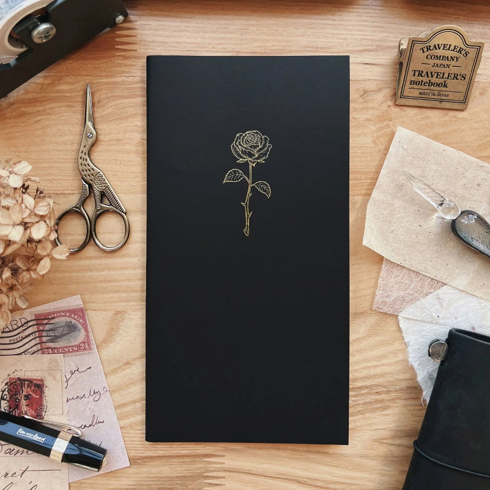 Black notebook with a rose design on a wooden surface with stationery items.